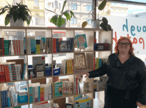 A counsellor stands in front of the large selection of books in the Rough Patch mental health shop. The counsellor has bright orange hair and is smiling. She is wearing black clothes and has glasses on.